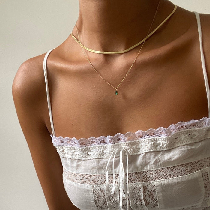 A person wears the Kismet Hailey Set, featuring a white lace-trimmed spaghetti strap top and two gold necklaces—a herringbone chain and an emerald green necklace—against a plain light background.