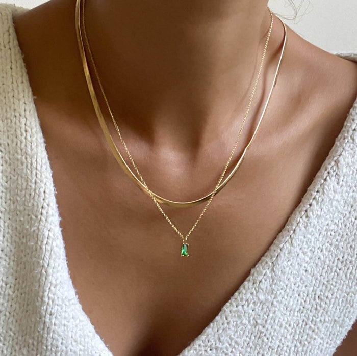 A close-up of a person wearing a white textured sweater and the Kismet Hailey Set by LLUME Jewelry, featuring three elegantly layered gold necklaces, including a herringbone chain and an emerald green necklace.