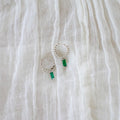 The Kismet Hoops Silver by LLUME Jewelry, featuring silver hoops adorned with small beads and emerald green rectangular gemstones, are displayed on a crinkled white fabric background.