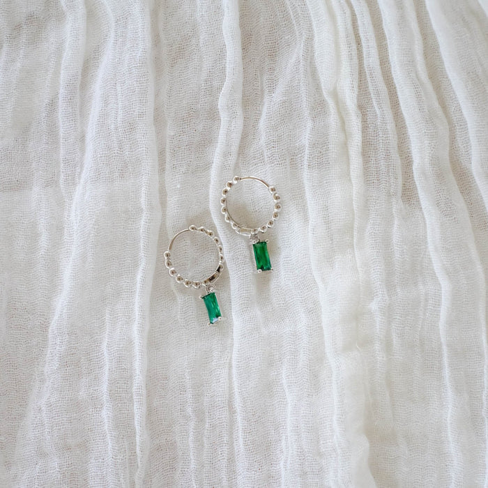 The Kismet Hoops Silver by LLUME Jewelry, featuring silver hoops adorned with small beads and emerald green rectangular gemstones, are displayed on a crinkled white fabric background.