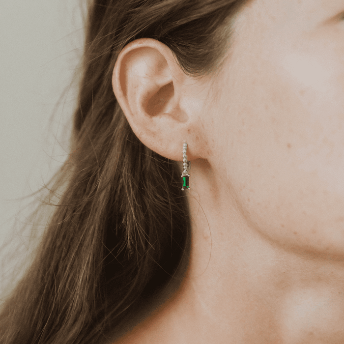 A close-up of a woman's ear adorned with LLUME Jewelry's Kismet Hoops Silver, featuring a green rectangular gemstone. Her brown hair is tucked behind her ear, showcasing these dainty silver earrings against a neutral background.