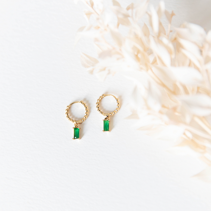 The Kismet Hoops by LLUME Jewelry—gold plated earrings with rectangular green gemstones—are displayed on a white surface, beside softly blurred beige dried foliage.