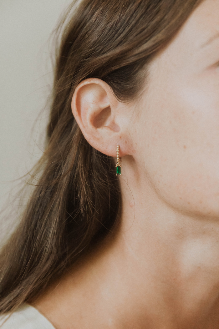 A close-up shows a woman's ear adorned with LLUME Jewelry's Kismet Hoops—gold plated earrings featuring a small, rectangular green gemstone. Her brown hair is tucked back to showcase the elegant design against her smooth, light-toned skin.