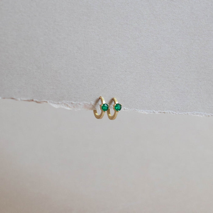 The Kismet Huggies by LLUME Jewelry are gold-plated green earrings with stone accents, shown on torn white paper against a minimalist, neutral backdrop.