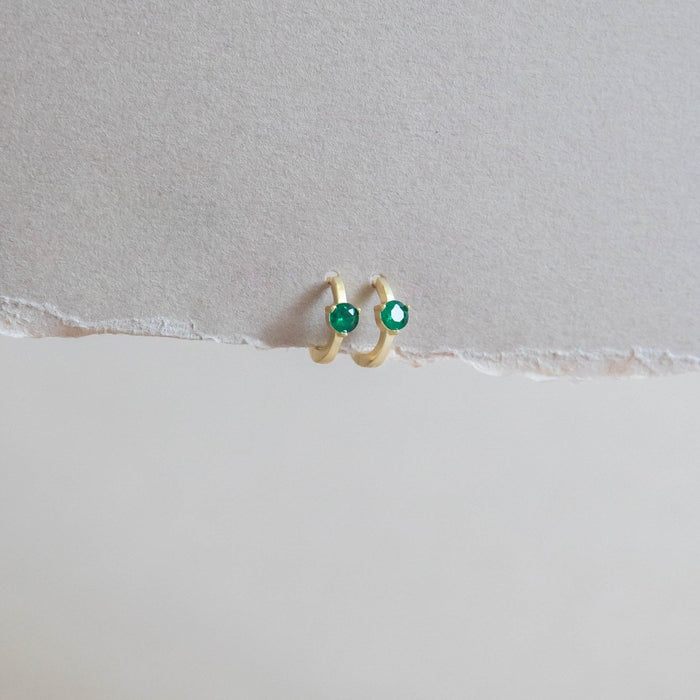 The Kismet Huggies by LLUME Jewelry are small gold-plated green gemstone earrings, shown against a textured, torn-edge light beige paper background.