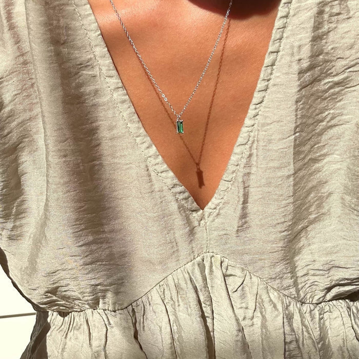 A close-up of a person in a light beige, textured V-neck dress, wearing LLUME Jewelry’s Kismet Necklace Silver—a dainty sterling silver piece with a small emerald green pendant. Sunlight casts soft shadows on the fabric and skin.