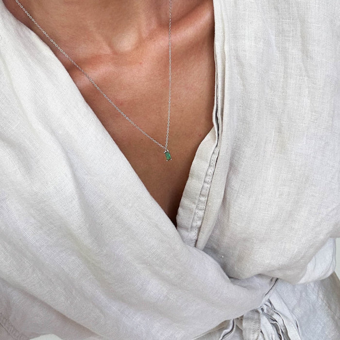 A person in a light linen wrap dress with a v-neck wears dainty jewelry—a LLUME Jewelry Kismet Necklace Silver featuring a small, rectangular emerald green pendant.