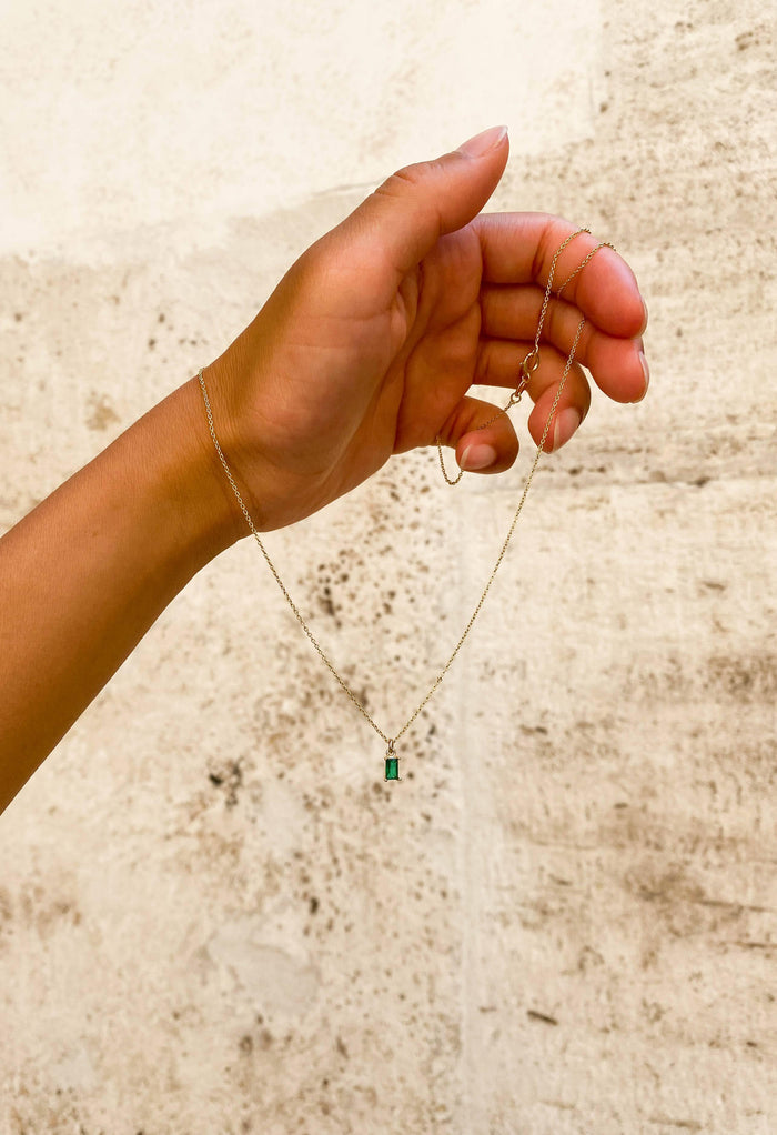 A hand displays the Kismet Necklace, featuring a delicate gold plated chain and a small emerald green charm pendant, set against a textured light background.