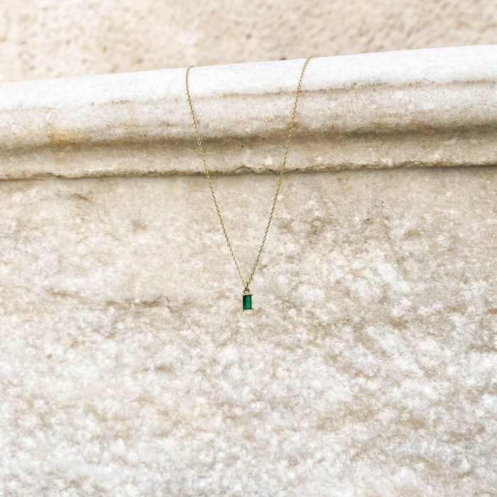 The Kismet Necklace features a small emerald green charm on a gold-plated chain, elegantly displayed against a textured, light stone background.