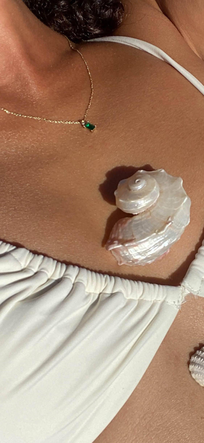 A person wearing a white top and the Kismet Necklace—a gold-plated chain with an emerald green charm—has a shiny white seashell on their chest, sunlight casting gentle shadows across their skin.