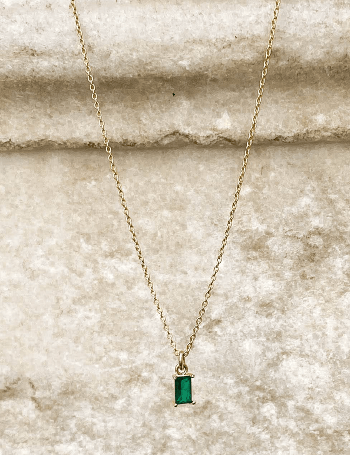 The Kismet Necklace features a gold plated chain with a small rectangular emerald green charm, showcased on a textured beige stone background.