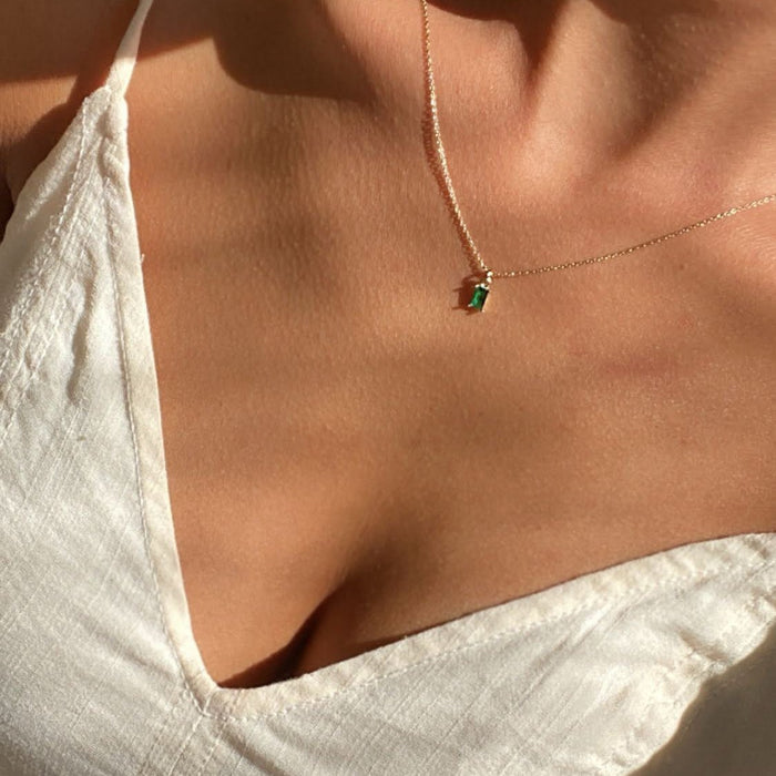 A close-up shows a person in a white textured V-neck top, wearing the Kismet Necklace with an emerald green charm, highlighted by soft natural light.