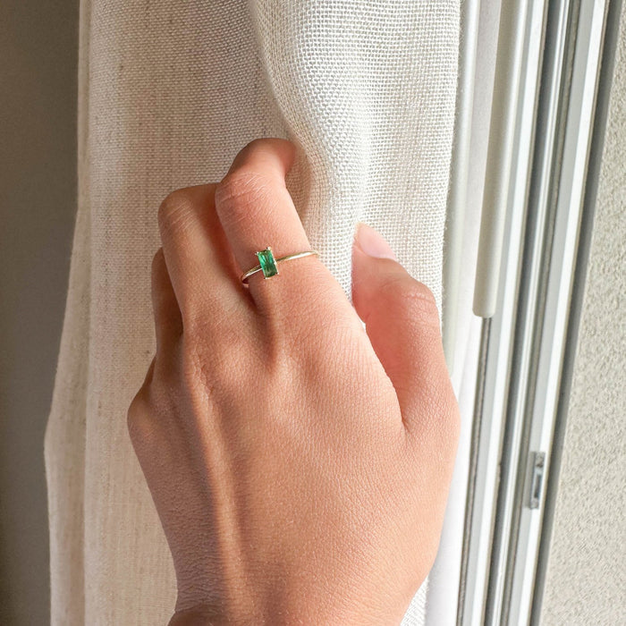 A hand with the LLUME Jewelry Kismet Ring, 14k gold-filled and set with an emerald green stone, rests against a light curtain near a window.
