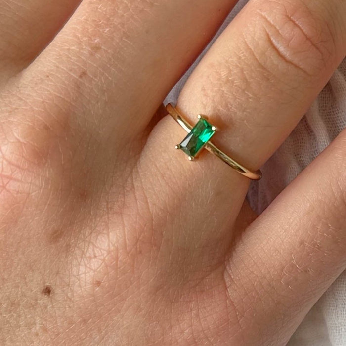 A hand wears LLUME Jewelry’s Kismet Ring—a thin 14k gold-filled band with a rectangular emerald green stone—set against soft white fabric.