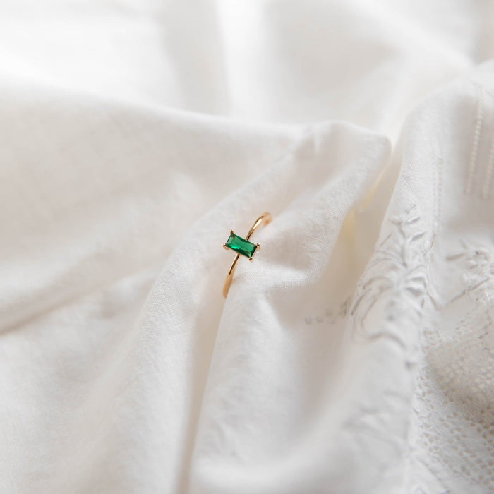 The Kismet Ring by LLUME Jewelry, featuring a rectangular emerald green stone and crafted in 14k gold-filled metal, is displayed on soft white embroidered fabric.