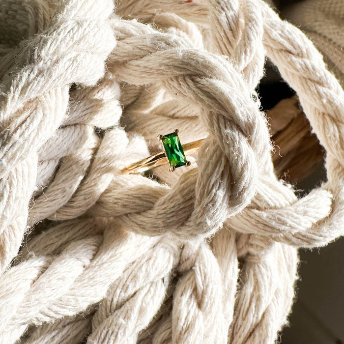 The Kismet Ring by LLUME Jewelry features a rectangular emerald green stone set in 14k gold-fill, shown atop thick cream-colored rope with sunlight highlighting its textured elegance—a striking piece from the Kismet Collection.