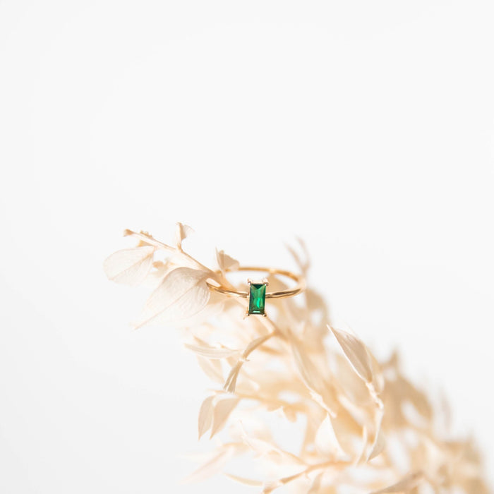 The Kismet Ring by LLUME Jewelry features a rectangular emerald green stone set in delicate 14k gold-filled metal, displayed on pale dried foliage against a white background.
