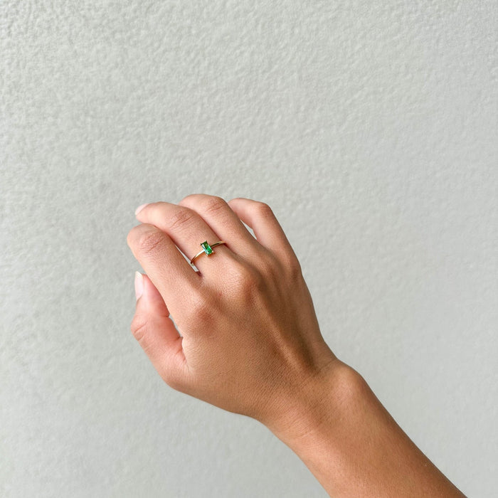 A hand with a light brown complexion wears the LLUME Jewelry Kismet Ring, a delicate 14k gold-filled piece with a small emerald green stone, shown against a plain light gray background.