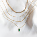 The Kismet Solstice Set by LLUME Jewelry displays five gold layered necklaces on white fabric, featuring mixed chain styles and an emerald green pendant on the bottom necklace for an elegant touch.