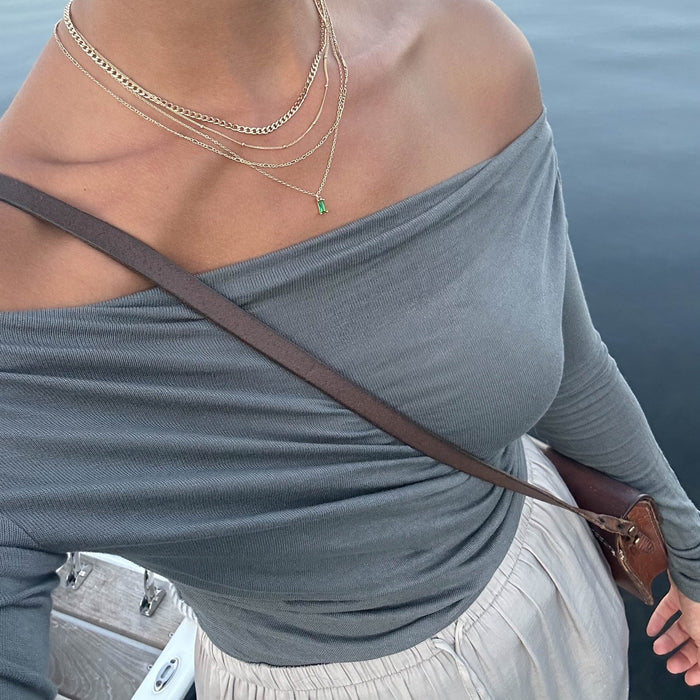 Wearing LLUME Jewelry’s Kismet Solstice Set, a person in a gray off-the-shoulder top, cream skirt, and brown crossbody bag stands by the water with a dock and boat in the background.