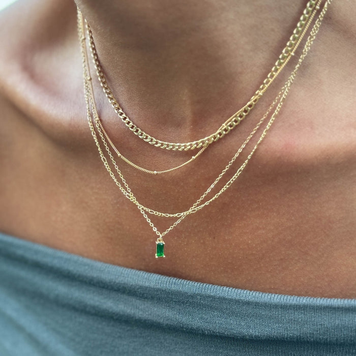 A close-up shows a person in a blue off-the-shoulder top wearing the LLUME Jewelry Kismet Solstice Set, a gold layered necklace set featuring an elegant emerald green pendant.