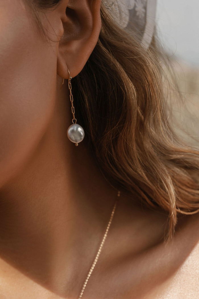 Close-up of a woman with light brown hair wearing LLUME Jewelry’s Lara Pearl Earrings—elegant gold-plated pearls. Part of a gold necklace appears on her neck, and the softly blurred background highlights the refined accessory.
