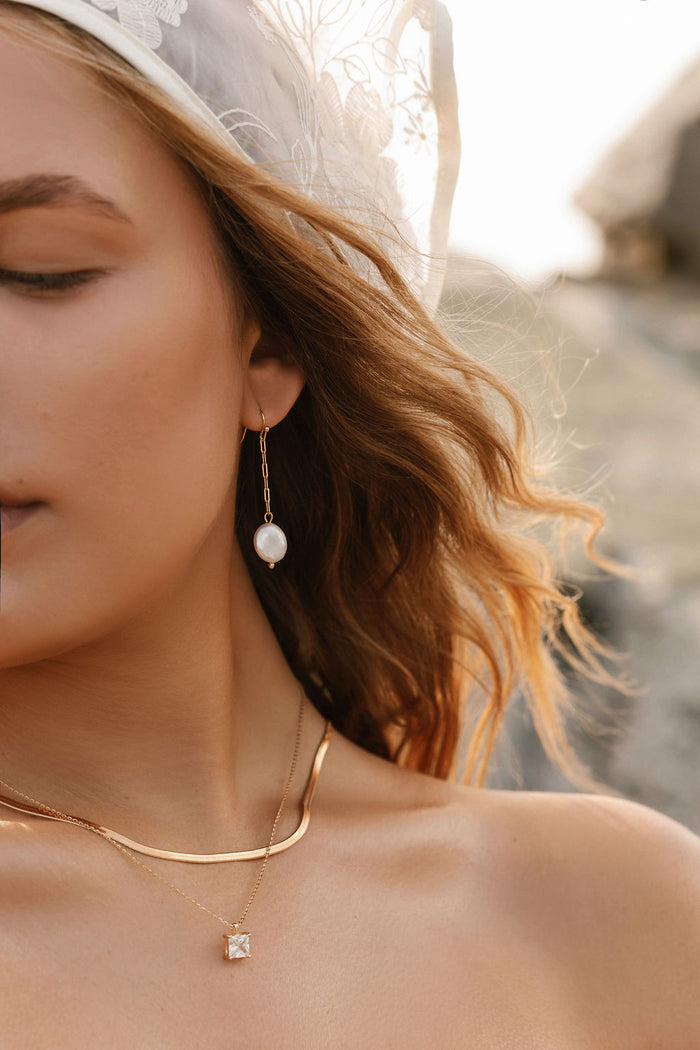 Close-up of a woman with wavy hair wearing the Lara Pearl Earrings by LLUME Jewelry, along with gold chain necklaces and a sheer, embroidered veil. Sunlight casts a warm glow, with softly blurred rocks in the background.