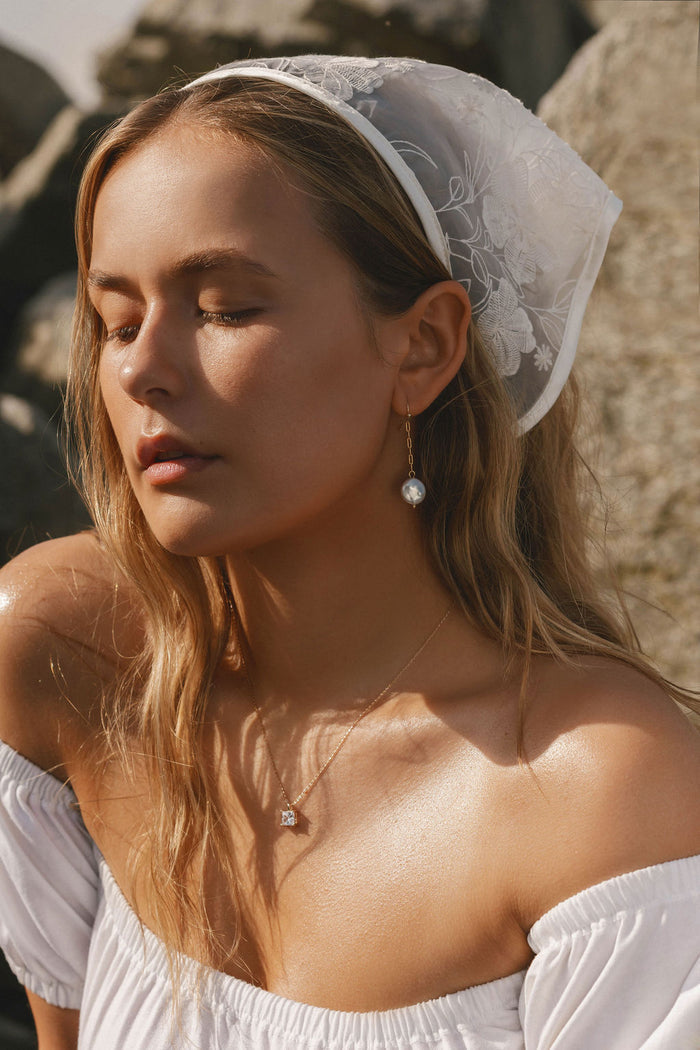 A young woman with closed eyes and long blonde hair wears LLUME Jewelry's Lara Pearl Earrings, a white lace headscarf, a delicate necklace, and an off-the-shoulder white top as she stands outdoors in the sunlight.
