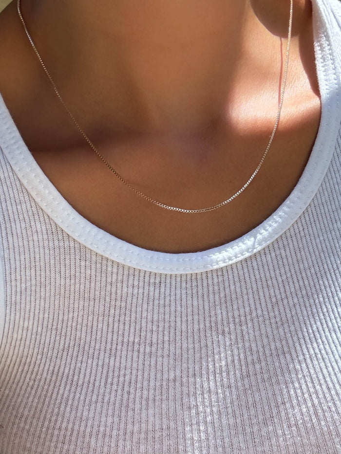 A close-up of a person wearing the LLUME Jewelry Lexi Box Chain necklace with a lobster clasp over a white ribbed tank top, sunlight casting gentle shadows on their skin.