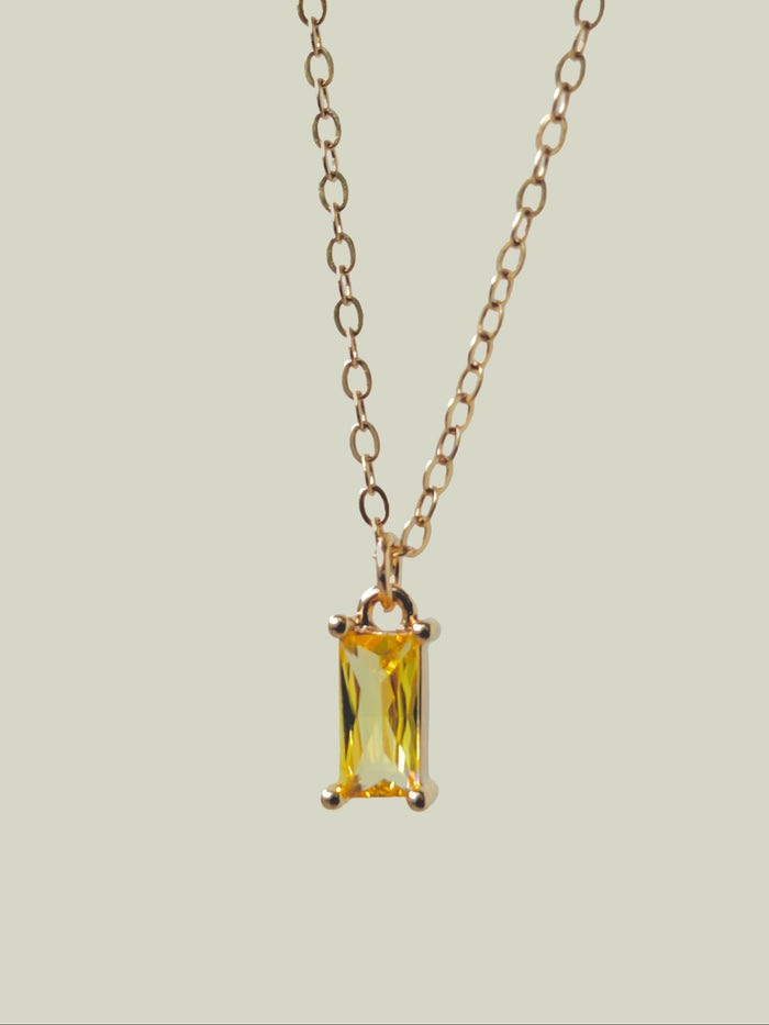 The Lila Necklace by LLUME Jewelry features a gold-plated chain and a rectangular yellow gemstone pendant in a minimalist gold frame, shown on a light beige background.