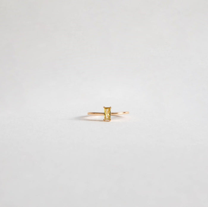 The Lila Ring by LLUME Jewelry features a minimalist 14k gold-filled band with a rectangular yellow gemstone, displayed against a light gray background.