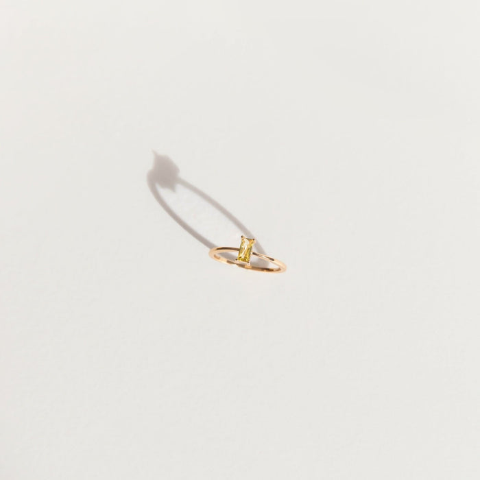 The Lila Ring by LLUME Jewelry features a rectangular yellow gemstone on a 14k gold-filled band, casting a soft shadow on a light surface.