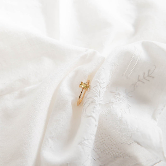 The Lila Ring by LLUME Jewelry—a 14k gold-filled stackable piece featuring a rectangular gemstone—rests on soft, white embroidered fabric, bathed in natural light.