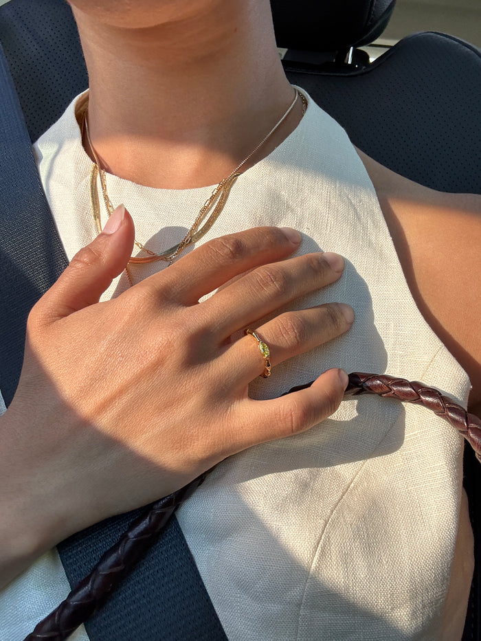 Wearing a cream halter top and layered gold necklaces, a person shows off the LLUME Jewelry Limon Ring—a lemon yellow accent—on their hand as they rest it on their chest, sunlight highlighting the ring and accessories while seated in a car with a seatbelt.