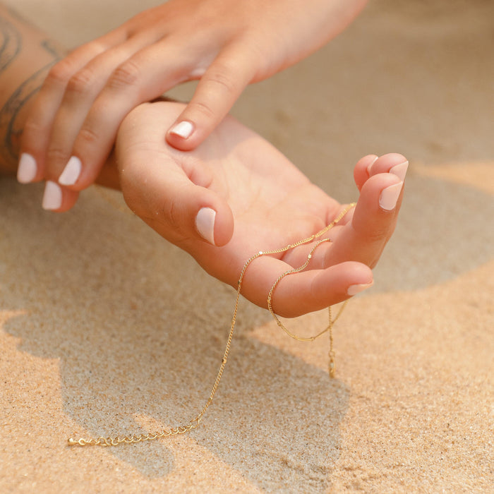 A close-up of hands with white manicured nails and a forearm tattoo rests on sandy ground, holding the Linden Chain necklace from LLUME Jewelry.