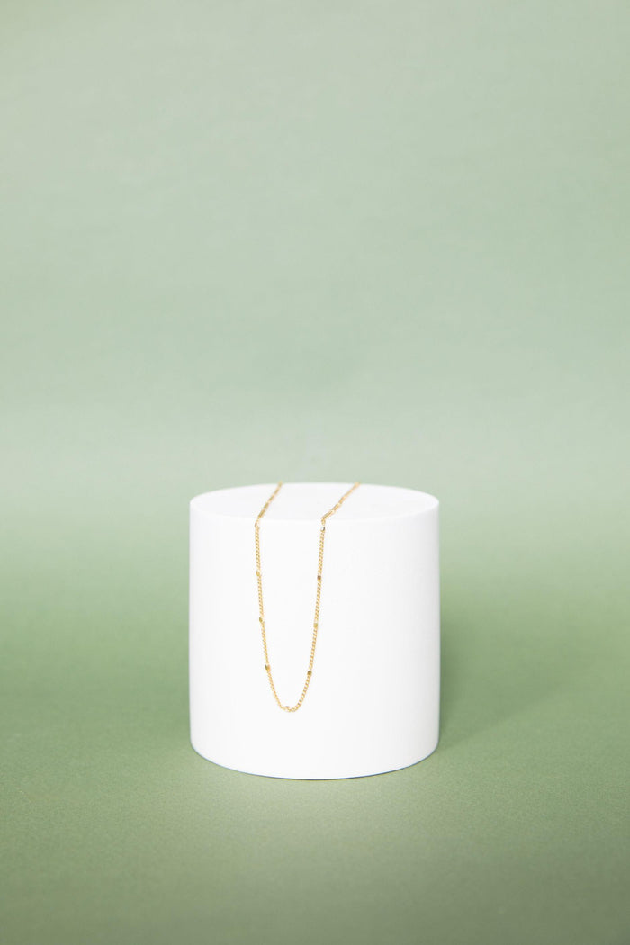 The Linden Chain by LLUME Jewelry, an 18K gold plated necklace, is displayed on a round white pedestal with a soft green backdrop—perfect for adding effortless elegance to any layered look.