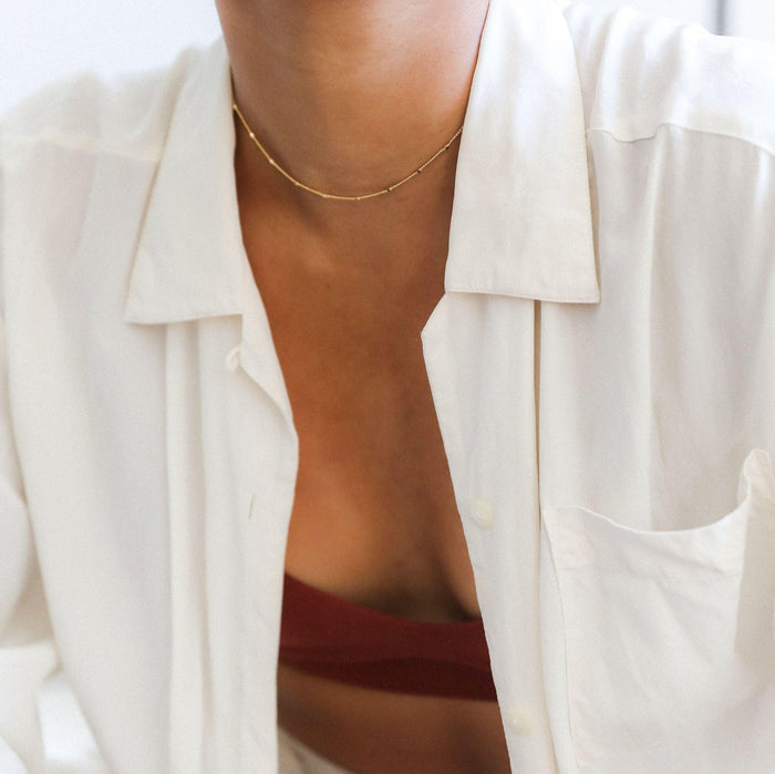 A person wears LLUME Jewelry's Linden Chain, styled with a rust top and white button-up. The photo focuses on their neck and chest, highlighting the subtle elegance of this dainty gold necklace layered for a chic look.