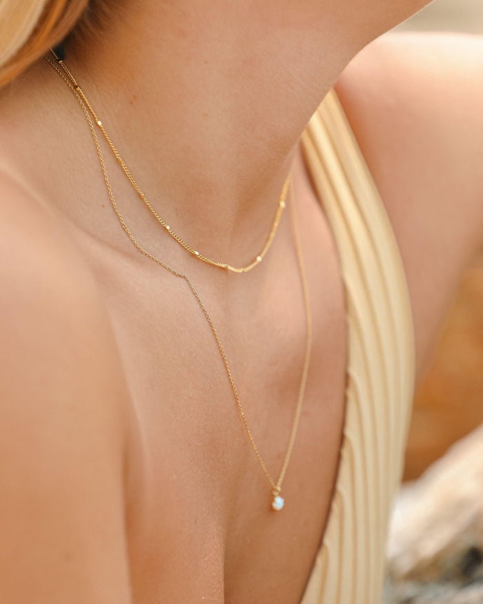 A close-up of a woman wearing LLUME Jewelry’s Linden Chain, an 18K gold plated layered necklace with a small pearl pendant, styled over a sleeveless beige top against a softly blurred background.