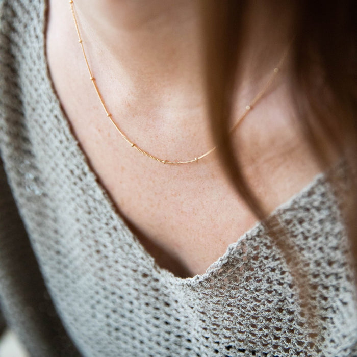 A close-up of a person wearing LLUME Jewelry's Linden Chain, a dainty gold necklace, with a textured grey knit top featuring a V-neckline. Brown hair softly frames the scene in the foreground.