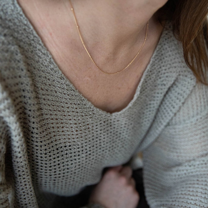 A close-up shows a person wearing a loose-knit light gray sweater and the LLUME Jewelry Linden Chain, an 18K gold plated necklace resting delicately on their neck, with their face hidden and hair softly draped to one side.