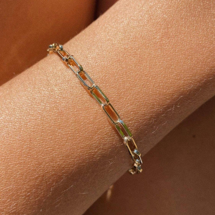 A close-up of the LLUME Jewelry Link Chain Bracelet on a person’s wrist, showcasing elongated rectangular links in a delicate gold-plated minimalist design against light brown skin.
