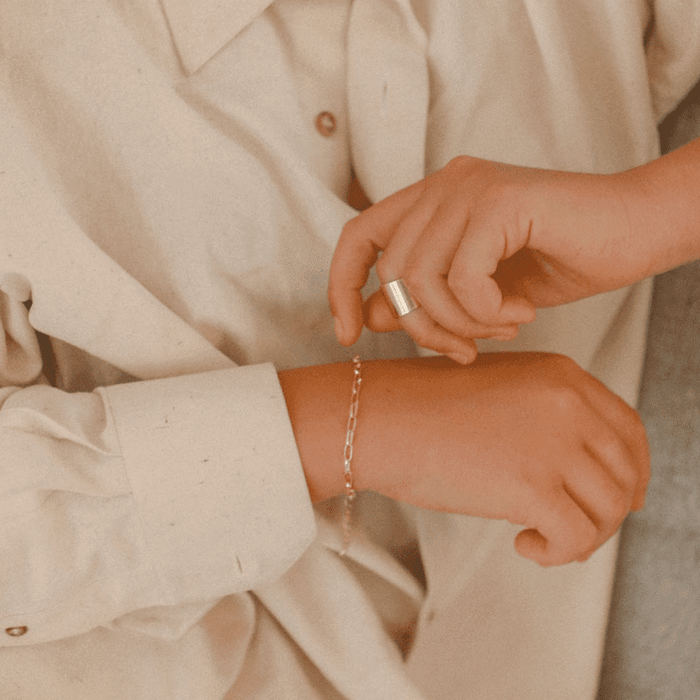 A person in a cream shirt fastens LLUME Jewelry's Link Chain Bracelet, a delicate gold-plated piece, on their wrist and also wears a large silver ring on one finger.
