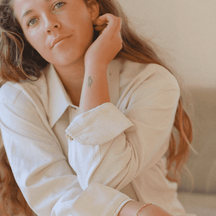 A person with long, wavy brown hair in a light button-up shirt gently touches their ear while looking at the camera. A small tattoo and LLUME Jewelry’s gold-plated Link Chain Bracelet decorate their wrist.