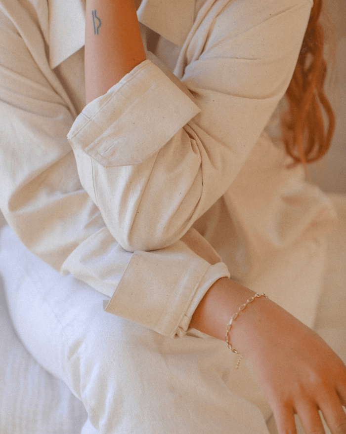 A person in a cream long-sleeve shirt and pants sits with one arm on their knee, showing a small tattoo and the LLUME Jewelry Link Chain Bracelet, an elegant gold-plated accessory, on their wrist.
