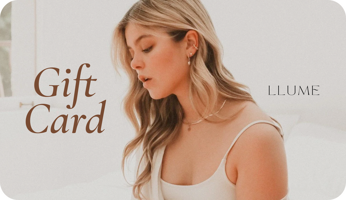 A blonde woman in a white top sits on a bed, gazing down thoughtfully. Beside her, “LLUME Jewelry Gift Card” appears in elegant font. This digital gift card from LLUME Jewelry has no expiry and is set against soft, neutral tones.
