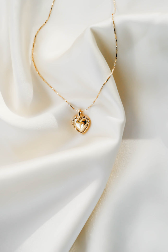 The Love Heart Necklace by LLUME Jewelry features a gold-plated heart pendant with a smaller inner heart, creating an elegant and romantic look as it rests on soft, white, silky fabric.