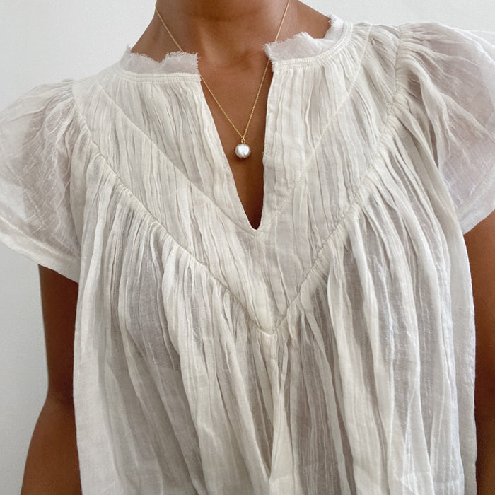A person wears a white, pleated, semi-sheer blouse with a frayed neckline and cap sleeves, accessorized with LLUME Jewelry's Lovegood Necklace—a gold vermeil chain featuring a single pearl pendant.