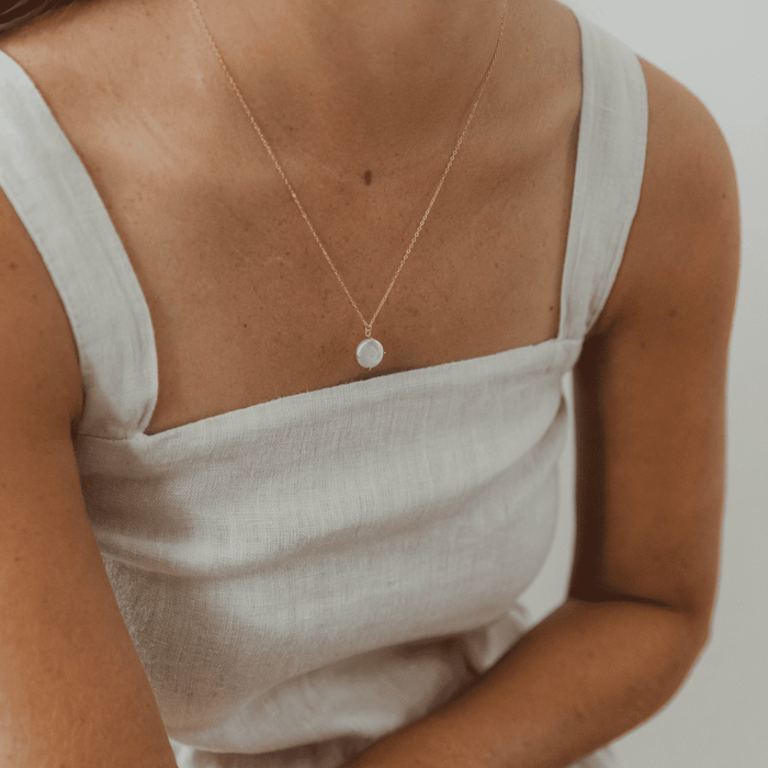 A woman wears a sleeveless, square-neck, light linen dress and the LLUME Jewelry Lovegood Necklace, a delicate gold vermeil chain with a round translucent pendant. The background is plain and softly lit.