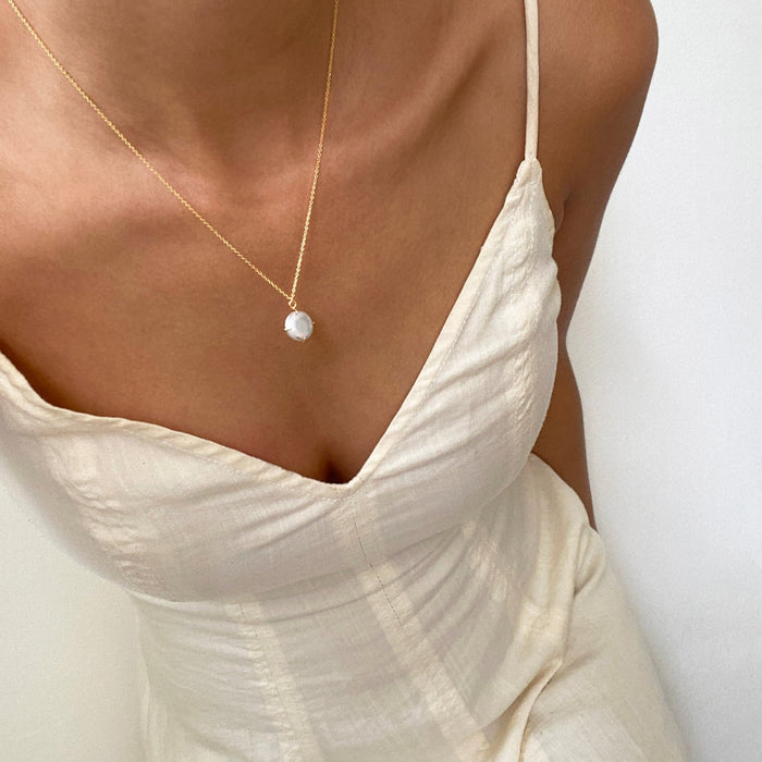 A person wears a cream spaghetti strap dress and the LLUME Jewelry Lovegood Necklace, featuring a round gold vermeil pendant, against a plain light background.