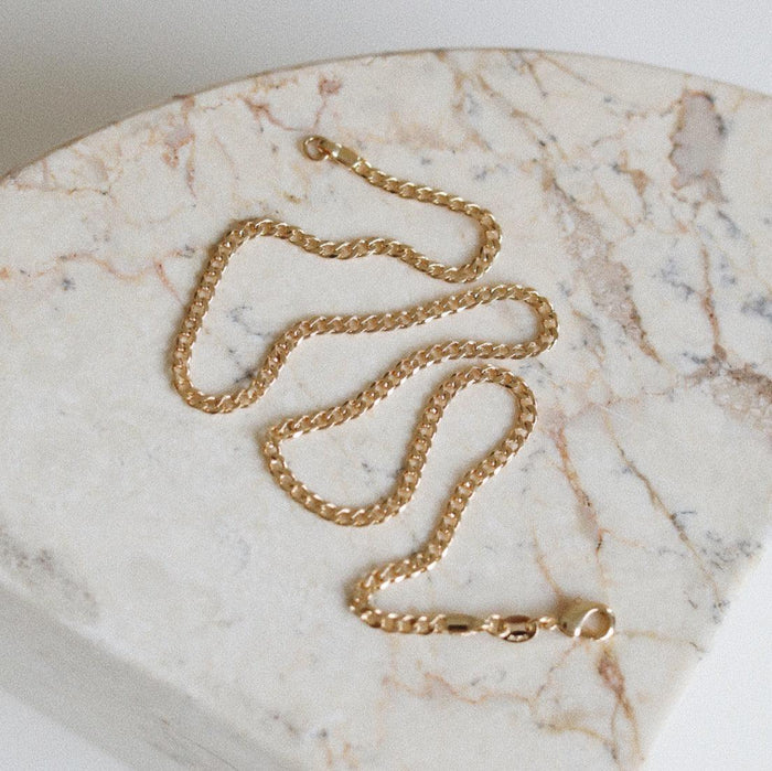 The LLUME Jewelry Marie Chain, an 18k gold-filled necklace with a lobster clasp, is displayed in wavy lines on a light marble surface featuring beige and tan veining.